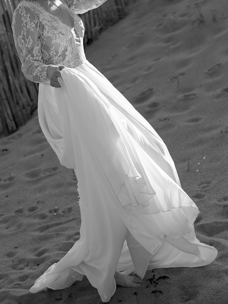 New England Coastal Elopement | Sincerely Liz Photography Vermont Wedding Photographer | Bride in a flowing white dress with lace sleeves walks on a sandy beach. The sun casts soft shadows, creating a serene and romantic atmosphere.
