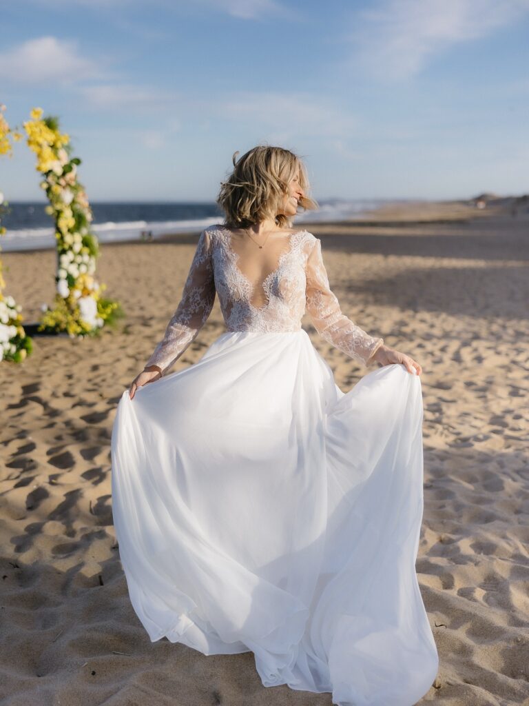 New England Coastal Elopement | Sincerely Liz Photography Vermont Wedding Photographer | A bride in a lace wedding dress stands on a beach, holding her skirt. The background shows a floral arch and the ocean under a clear blue sky.