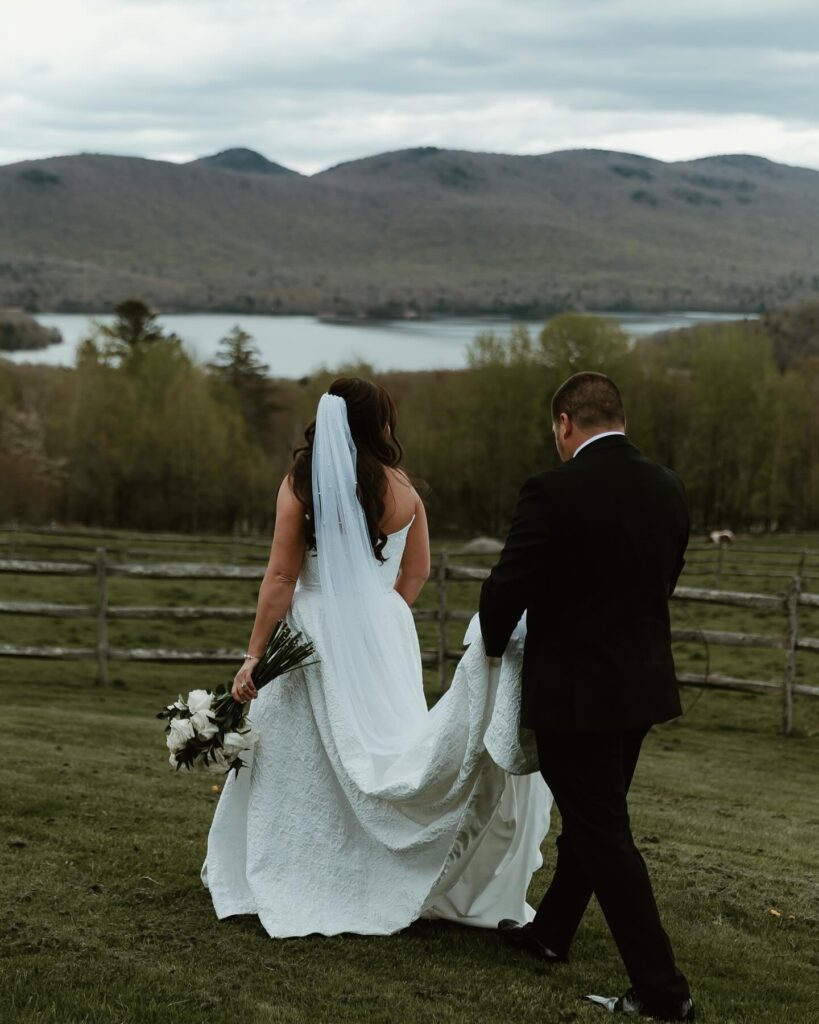Vermont Wedding Venues | Sincerely Liz Photography Vermont Wedding Photographer | Bride and groom walking on grass with mountains and lake in the background. The bride holds a bouquet and veil, creating a serene, romantic atmosphere.