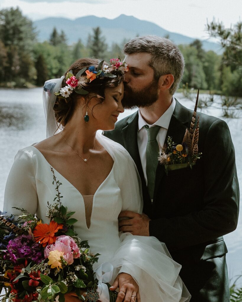 The groom kisses the bride’s forehead beside a mountain lake, a tender moment from a Vermont mountain wedding.