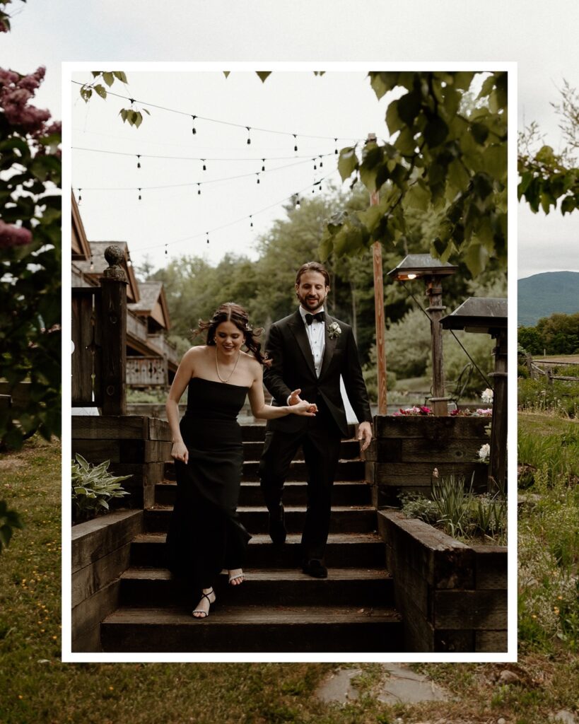 Newlyweds in black formal attire walking down rustic wooden steps beneath string lights, an elegant setting for a Von Trapp Lodge wedding in Vermont.