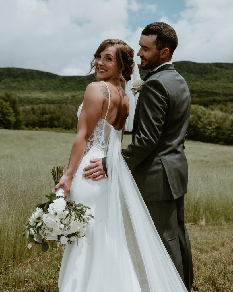 Vermont Wedding Venues | Sincerely Liz Photography Vermont Wedding Photographer | A joyful bride and groom stand embracing in a lush green field. The bride holds a bouquet of white flowers, her smile radiant against a backdrop of rolling hills and clouds.