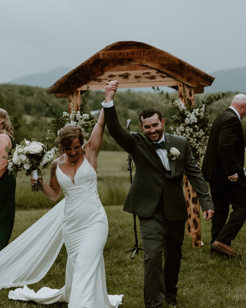 Vermont Wedding Venues | Sincerely Liz Photography Vermont Wedding Photographer | A joyful newlywed couple walks hand in hand under a rustic wooden arch adorned with flowers, smiling widely against a serene, natural backdrop.
