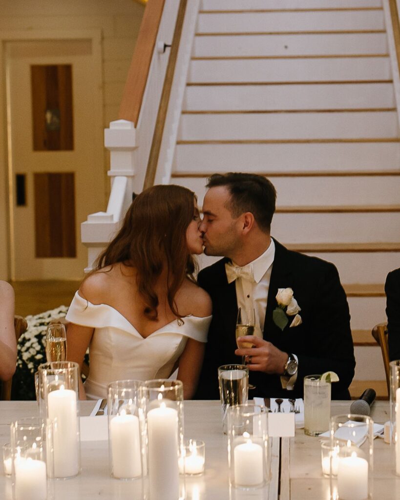 Vermont Wedding Venues | Sincerely Liz Photography Vermont Wedding Photographer | A bride in an off-the-shoulder dress and groom in a tuxedo lovingly kiss at a table adorned with lit candles, creating a warm, romantic ambiance.