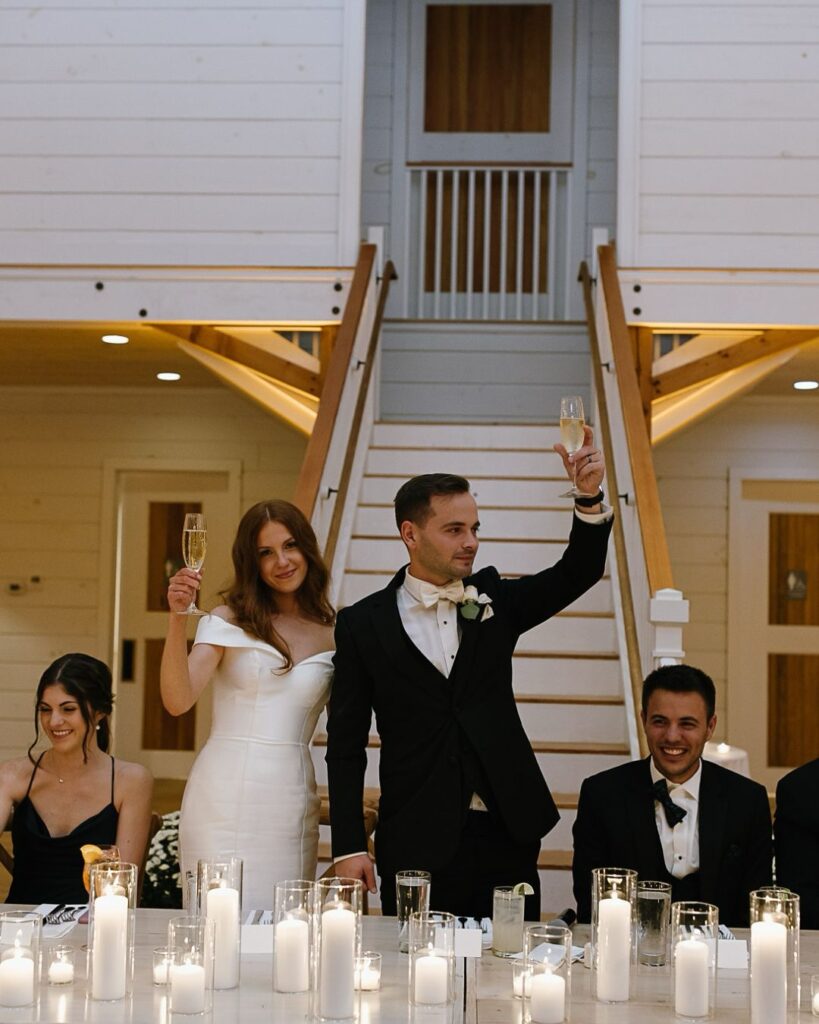 Vermont Wedding Venues | Sincerely Liz Photography Vermont Wedding Photographer | A bride and groom stand joyfully at a candlelit reception table, raising champagne glasses. Two seated guests beside them smile warmly, set against a wooden staircase backdrop.