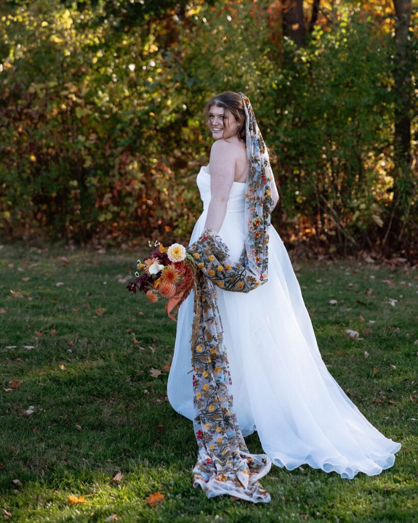 Vermont Wedding Venues | Sincerely Liz Photography Vermont Wedding Photographer | Bride in a white gown stands in a grassy field, smiling back. She holds a colorful bouquet with a floral scarf draped over her shoulder. Sunny, autumn setting.
