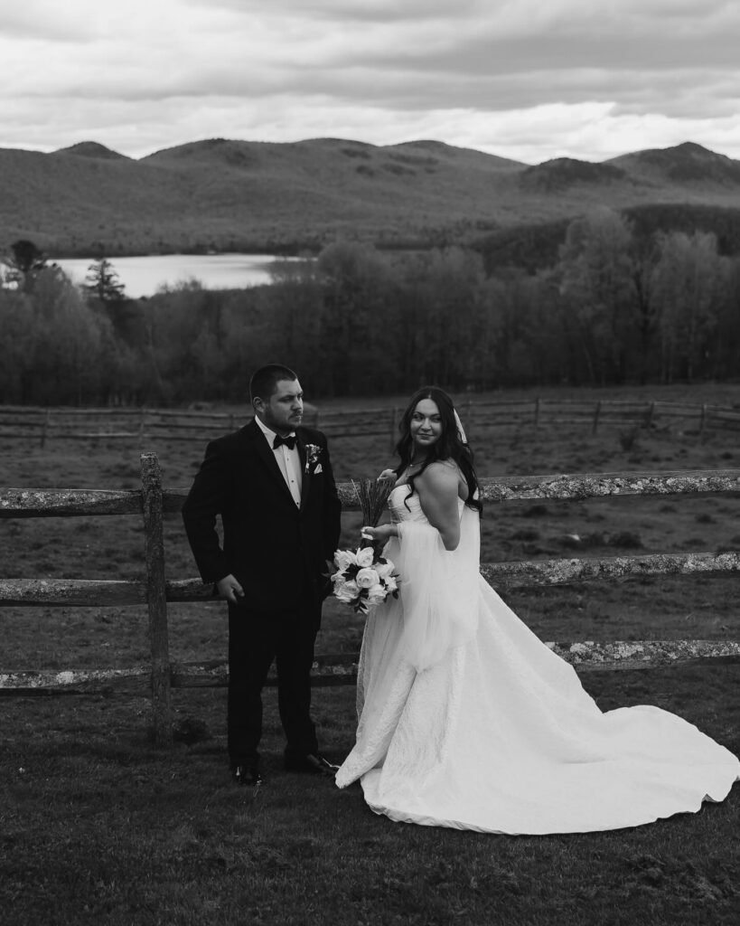 Vermont Wedding Venues | Sincerely Liz Photography Vermont Wedding Photographer | Bride and groom in formal attire pose by a wooden fence, with a serene lake and mountains in the background. The scene conveys romance and tranquility.