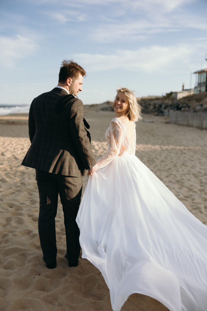 A newlywed couple walks hand in hand along a sandy beach, reflecting the relaxed spirit of a Vermont destination wedding.