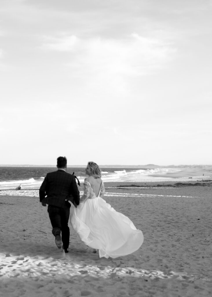Vermont Wedding Venues | Sincerely Liz Photography Documentary and Editorial Wedding Photography | A couple runs hand in hand on a sandy beach toward the ocean. The bride's dress flows in the breeze. The scene conveys joy and freedom.