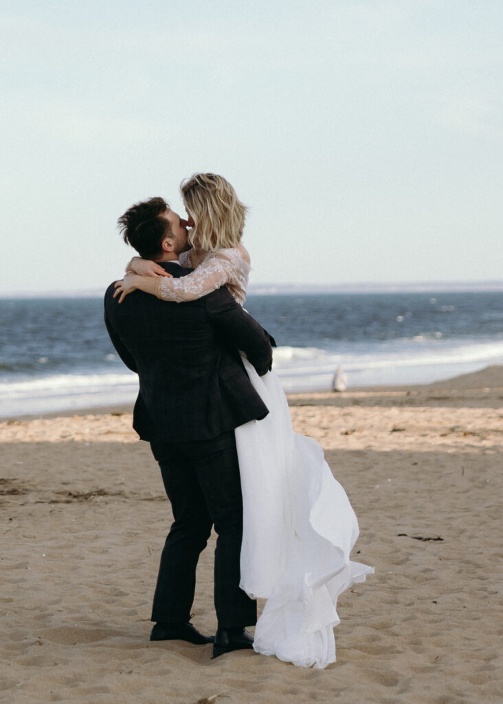 New England Coastal Elopement | Sincerely Liz Photography Vermont Wedding Photographer | A couple embraces joyfully on a sandy beach, with the ocean in the background. The woman wears a flowing white dress, and the man lifts her gently.