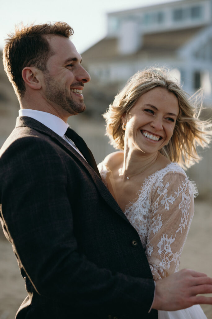 New England Coastal Elopement | Sincerely Liz Photography Vermont Wedding Photographer | A joyful couple embraces on a sunny beach. The woman in a lace wedding dress smiles radiantly, while the man in a dark suit laughs softly.