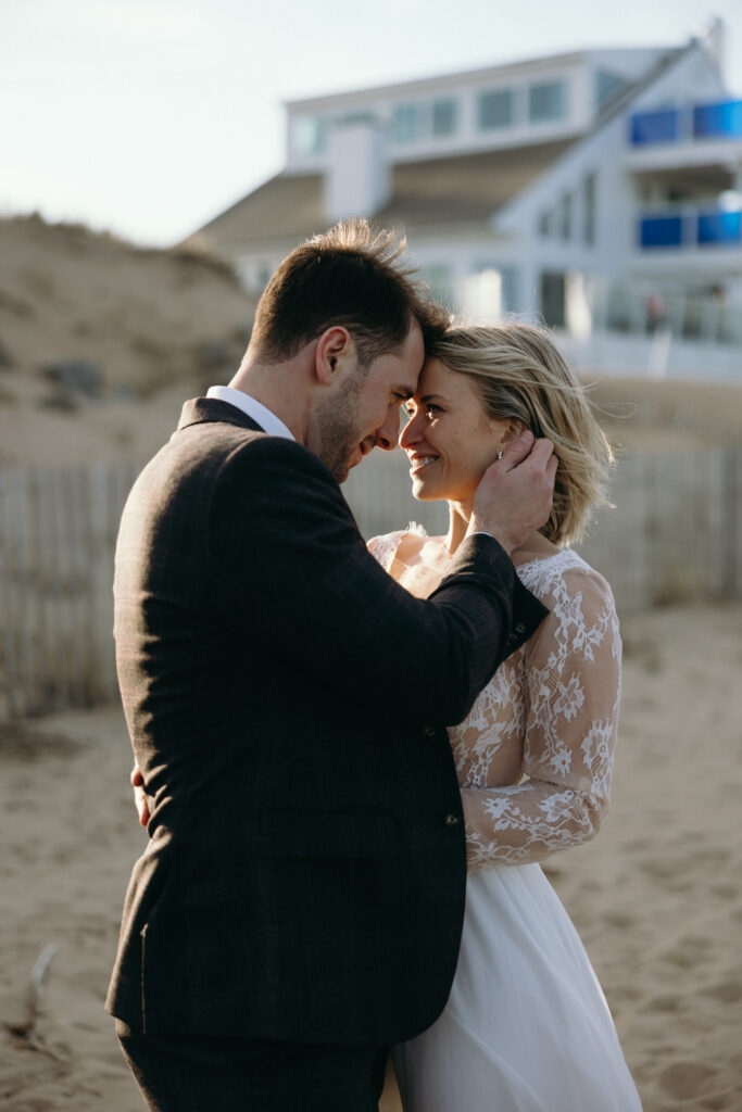 New England Coastal Elopement | Sincerely Liz Photography Vermont Wedding Photographer | A couple embraces on a beach, smiling warmly at each other. The woman wears a lace wedding gown, the man a dark suit. A building is visible in the background.