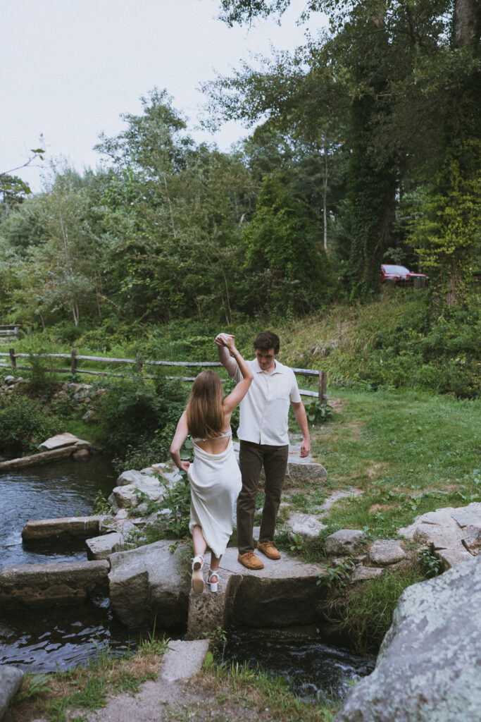 Engagement Vermont Photographer | Sincerely Liz Photography Vermont Wedding Photographer | A couple is outdoors, crossing a stream on large stones. The woman in a white dress balances as the man in a white polo supports her. Lush greenery surrounds them, creating a serene, romantic atmosphere.