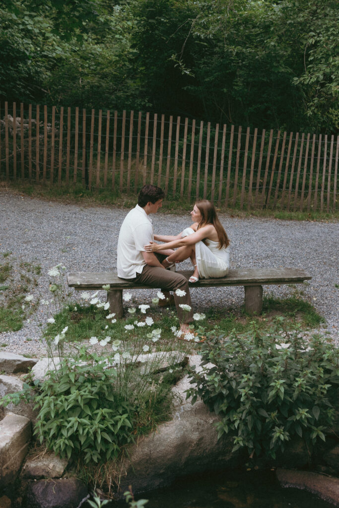 Engagement Vermont Photographer | Sincerely Liz Photography Vermont Wedding Photographer | A couple sits closely on a wooden bench in a peaceful, green garden. They're surrounded by blooming white flowers, exuding a serene and intimate mood.