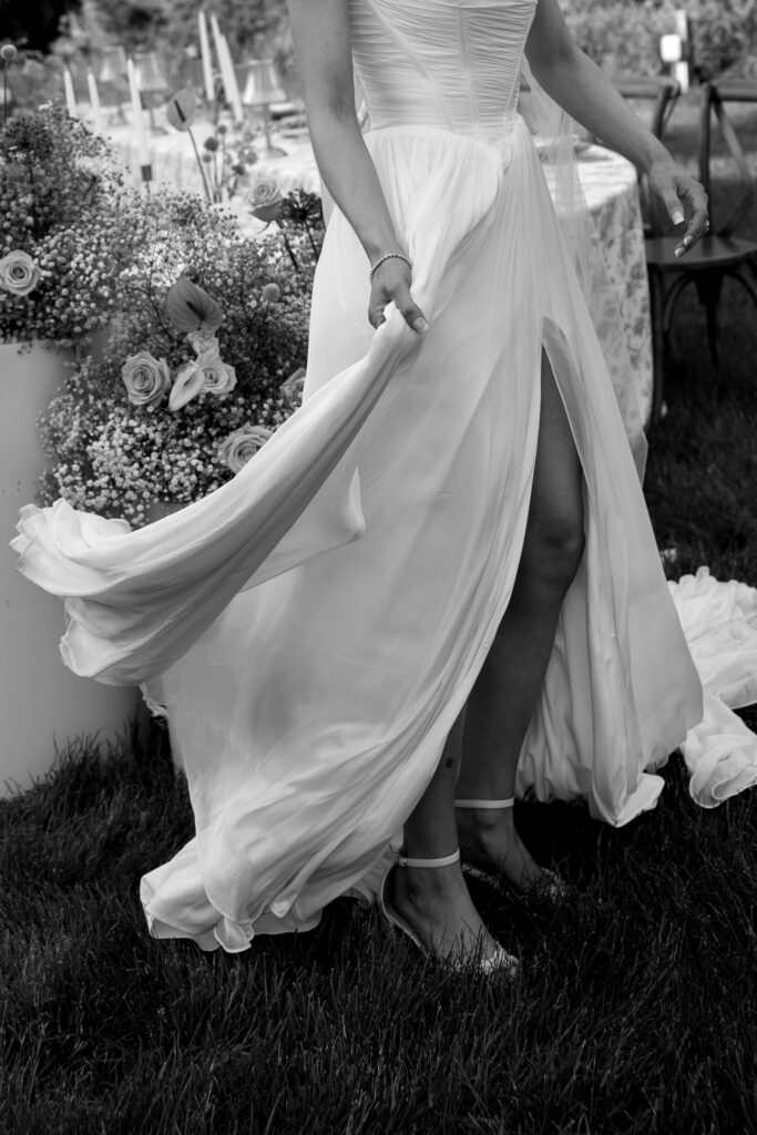 Black-and-white detail of a bride lifting her flowing gown beside soft garden florals, an editorial moment suited to Vermont inn wedding photography.