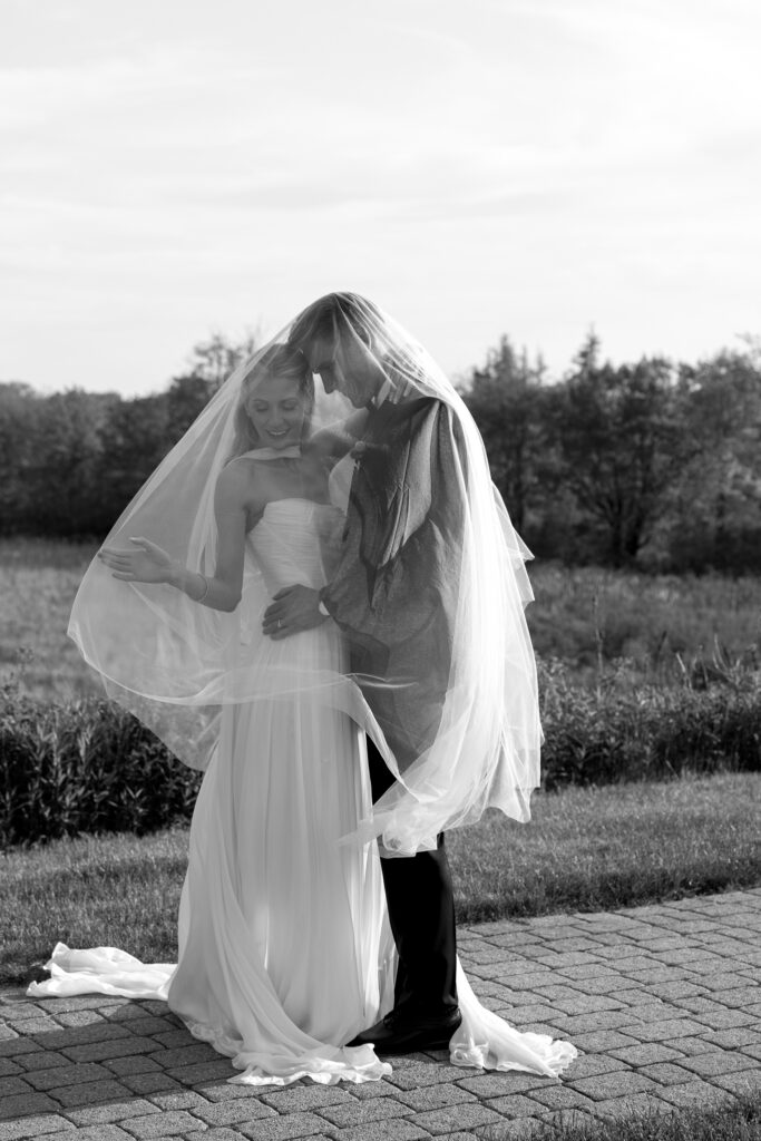 Black-and-white portrait of newlyweds embracing beneath the bride’s flowing veil outdoors, a quiet example of candid wedding photography in Vermont.