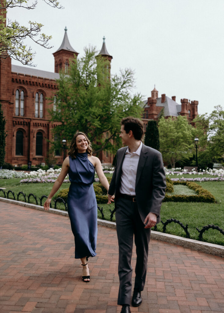 Engagement Vermont Photographer | Sincerely Liz Photography Vermont Wedding Photographer | A couple walks along a brick path, holding hands and smiling. They're in front of a historic building with towers, surrounded by greenery and flowers.