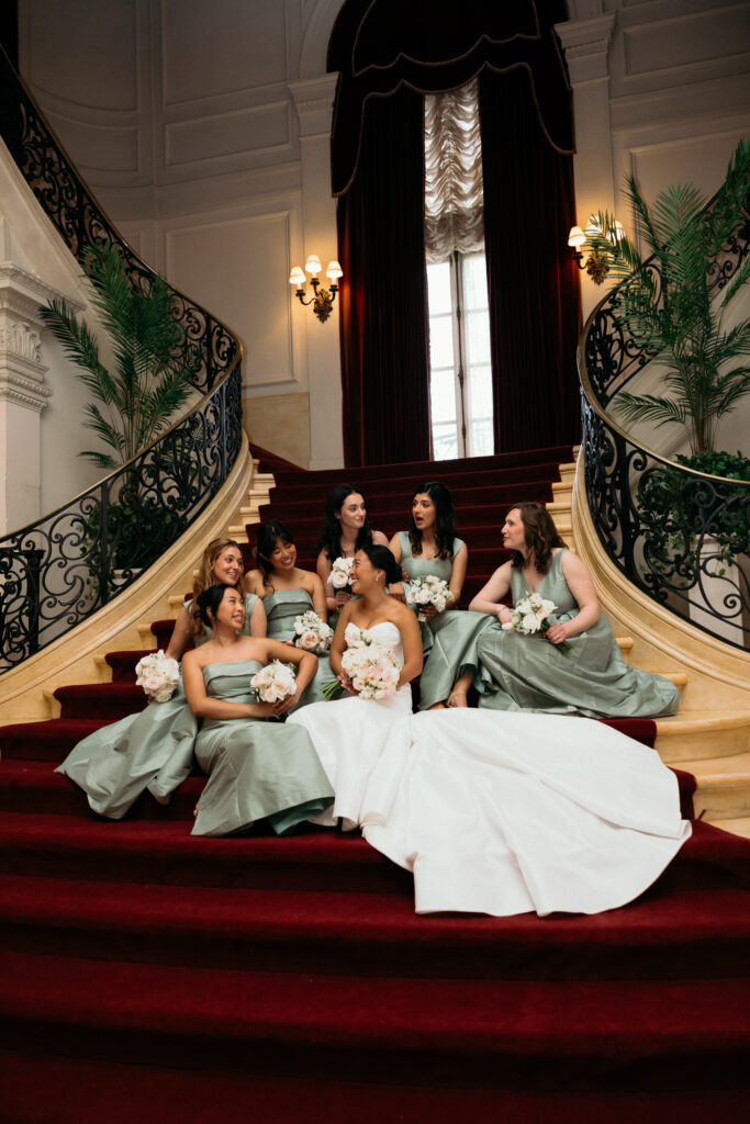 A bride sits with her bridesmaids on a grand curved staircase, capturing an elegant moment from a Vermont wedding celebration.