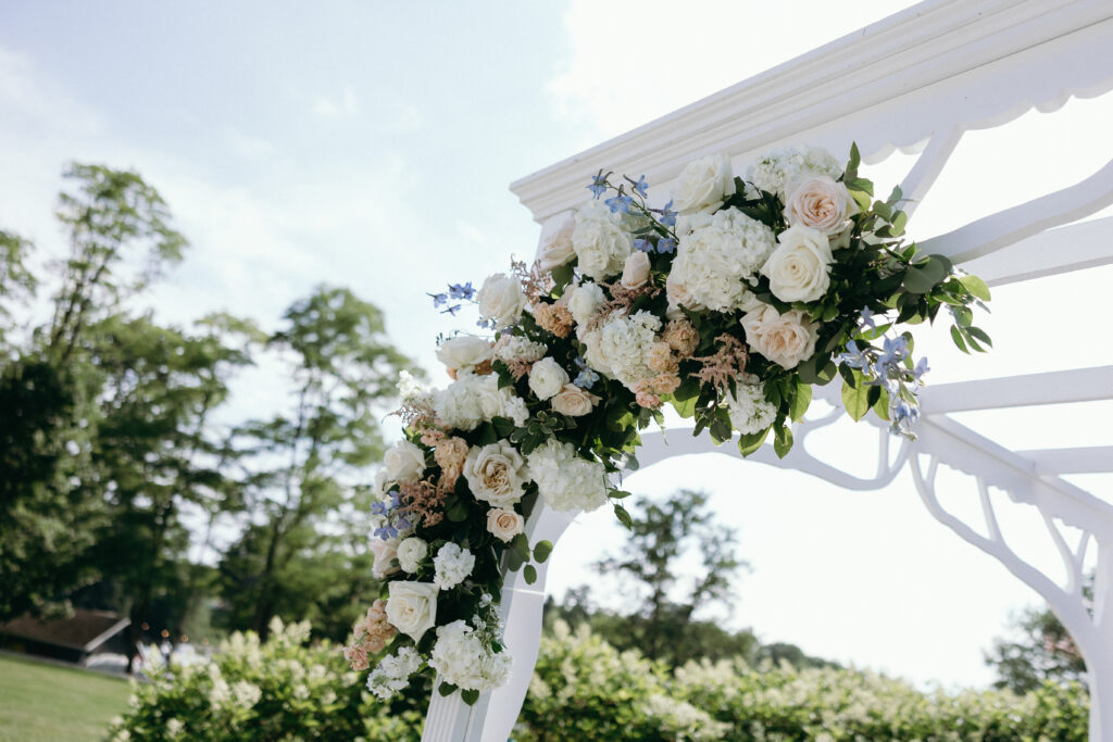 Vermont Wedding Venues | Sincerely Liz Photography Vermont Wedding Photographer | Elegant floral arrangement on a white wedding arch outdoors, featuring white, peach, and blush roses with green leaves. A serene, festive atmosphere.