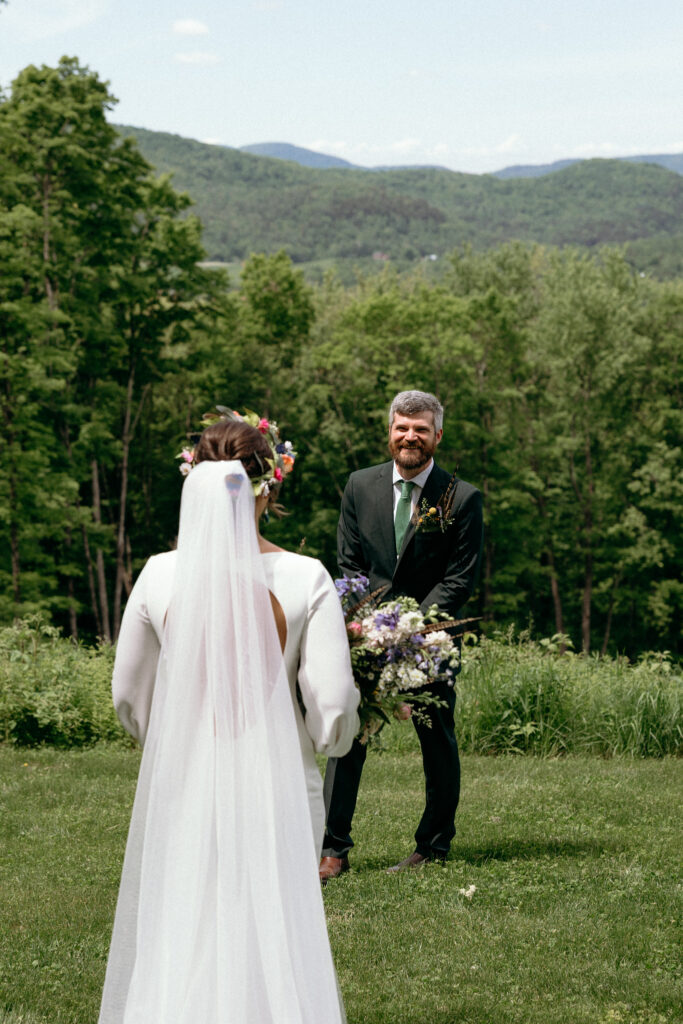 Vermont Wedding Venues | Sincerely Liz Photography Documentary and Editorial Wedding Photography | A bride in a white dress with a floral crown approaches a smiling groom holding a bouquet. They stand on a grassy field with trees and mountains in the background.