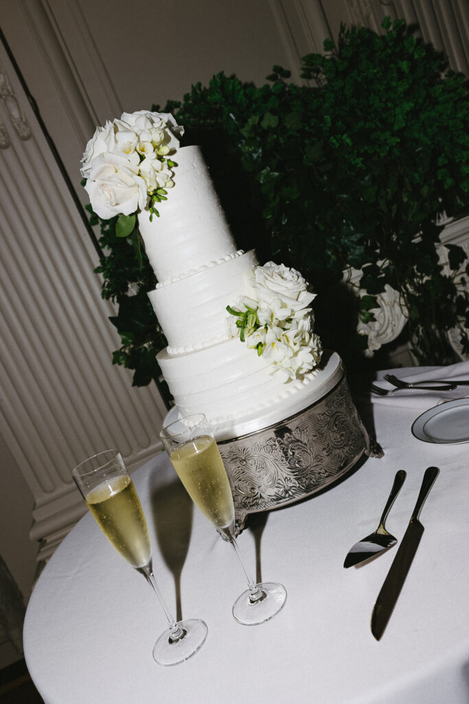 Tiered white wedding cake decorated with fresh flowers beside two champagne glasses, an elegant detail captured through true-to-tone wedding photography.
