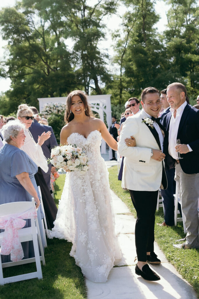 Vermont Wedding Photographer | Sincerely Liz Photography Documentary and Editorial Wedding Photography | A joyful bride in a white floral gown and a groom in a white tuxedo walk hand in hand down an outdoor wedding aisle surrounded by applauding guests.