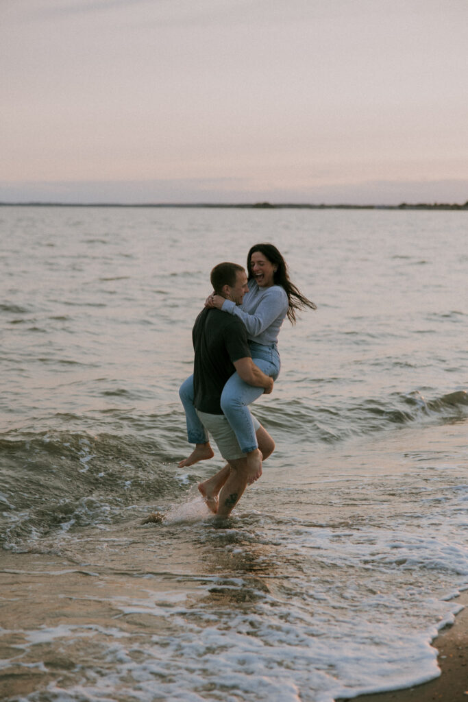 Engagement Vermont Photographer | Sincerely Liz Photography Vermont Wedding Photographer | Man carrying woman through gentle waves on a sandy beach at sunset. The couple is laughing, creating a playful and joyful atmosphere.