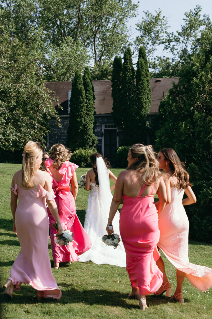 Vermont Wedding Photographer | Sincerely Liz Photography Documentary and Editorial Wedding Photography | Bridesmaids in pink dresses and the bride in a white gown with veil walk on a sunny lawn toward a stone building, exuding joy and anticipation.