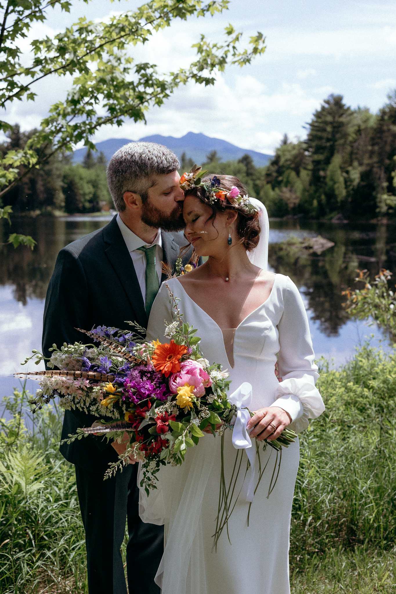 Vermont Wedding Venues | Sincerely Liz Photography Vermont Wedding Photographer | A bride and groom share an intimate moment by a serene lake. The bride holds a vibrant bouquet and wears a floral crown. The scene is peaceful and romantic.