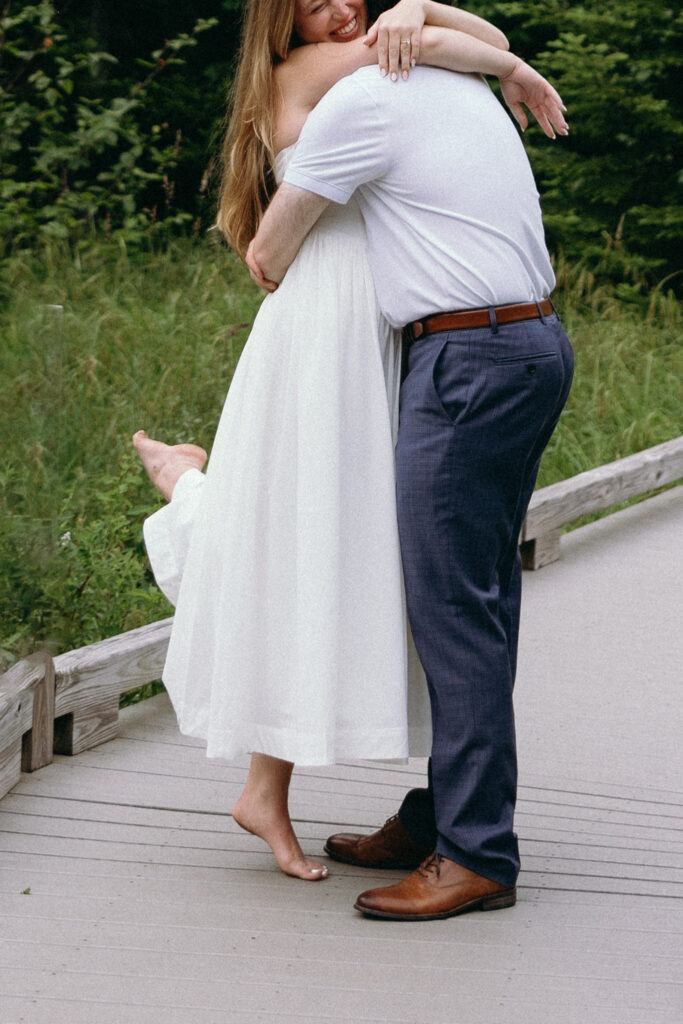 Engagement Vermont Photographer | Sincerely Liz Photography Vermont Wedding Photographer | A barefoot woman in a white dress is joyfully hugging a man in a white shirt and blue pants on a wooden path. Green foliage surrounds them.