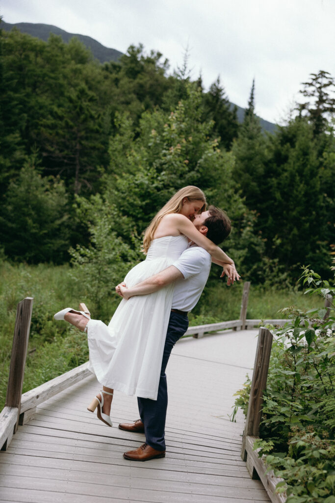 Engagement Vermont Photographer | Sincerely Liz Photography Vermont Wedding Photographer | A couple embraces happily on a wooden path surrounded by lush greenery. The woman is in a white dress, lifted by the man in casual attire. Romantic and joyful atmosphere.