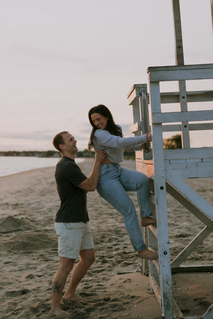 Engagement Vermont Photographer | Sincerely Liz Photography Vermont Wedding Photographer | A joyful couple on a sandy beach at sunset; one person helps the other climb a wooden lifeguard stand, both smiling and barefoot, creating a playful and warm atmosphere.