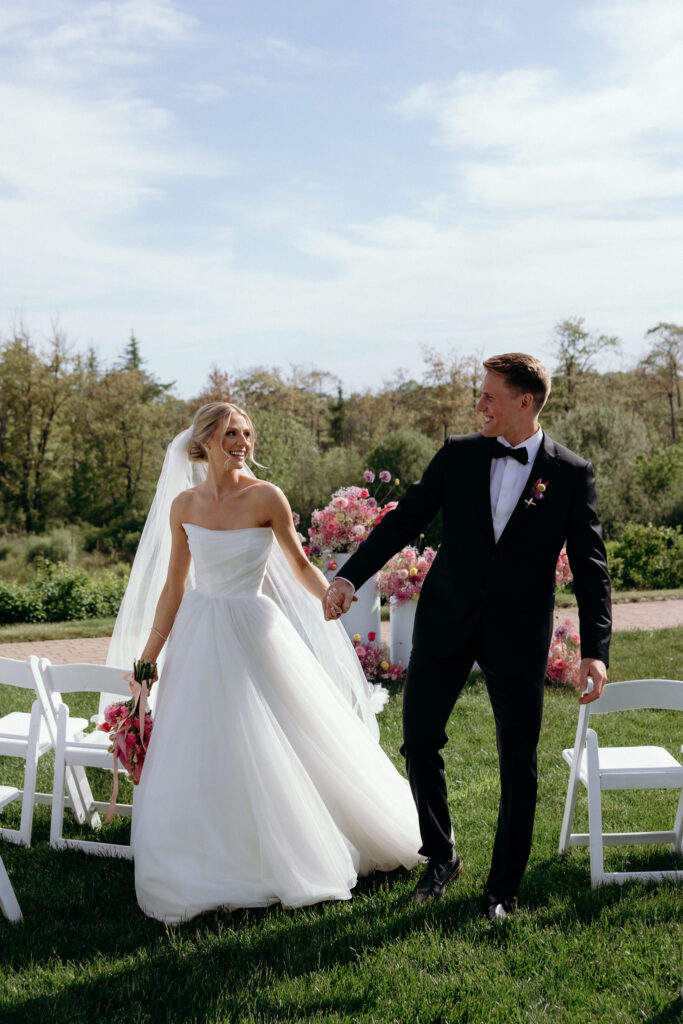 Newlyweds holding hands and smiling as they leave their outdoor ceremony, capturing the relaxed joy of a romantic inn wedding in Vermont.