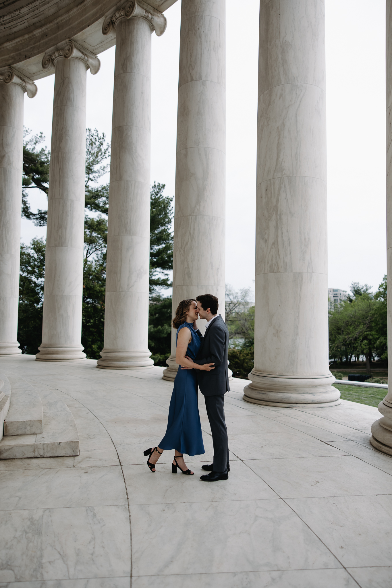 Engagement Vermont Photographer | Sincerely Liz Photography Vermont Wedding Photographer | A couple embraces romantically beneath tall, white columns, set on a marble floor. The woman wears a blue dress, the man a dark suit; trees are visible outdoors.