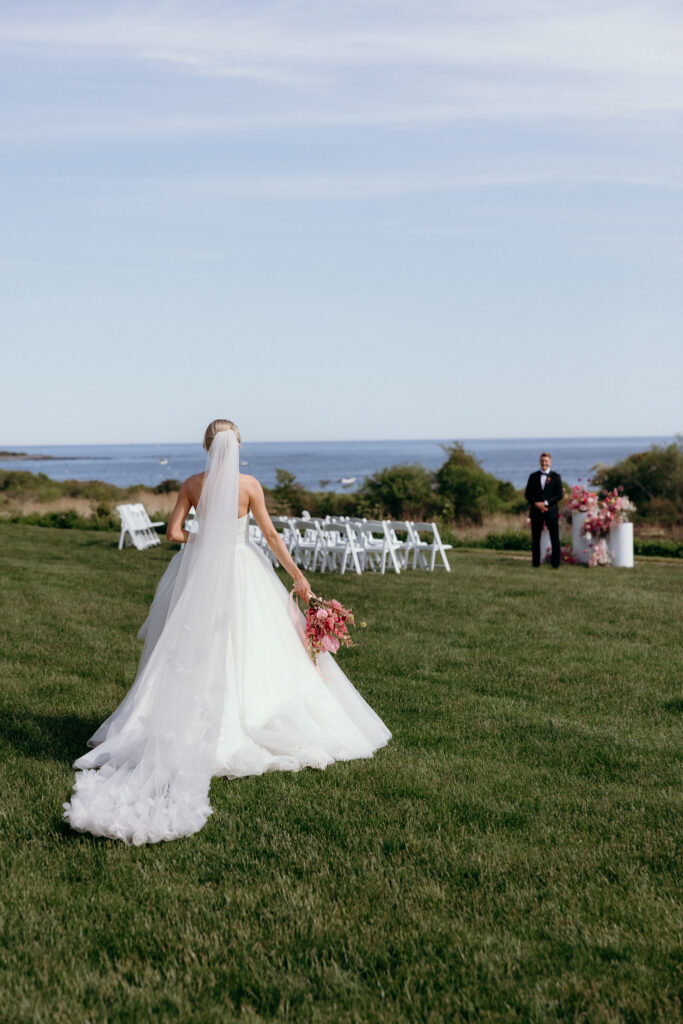 A bride walks across a grassy coastal lawn toward an outdoor ceremony setup with ocean views beyond, a beautiful fit for best wedding venues in Vermont by the water.