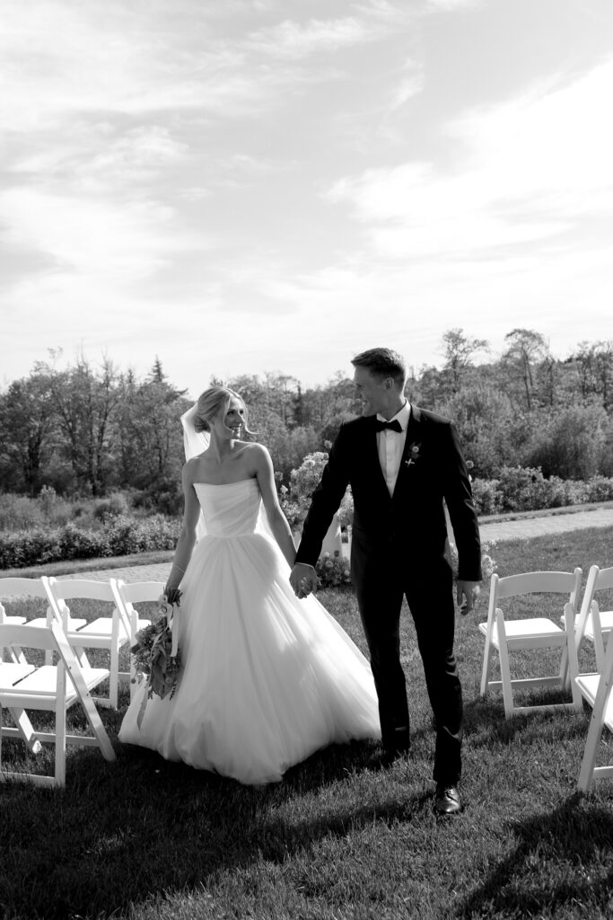 Vermont Wedding Photographer | Sincerely Liz Photography Documentary and Editorial Wedding Photography | A joyful couple walks hand-in-hand down a grassy aisle outdoors. The bride wears a flowing white gown, the groom a sharp suit. Empty white chairs are around.