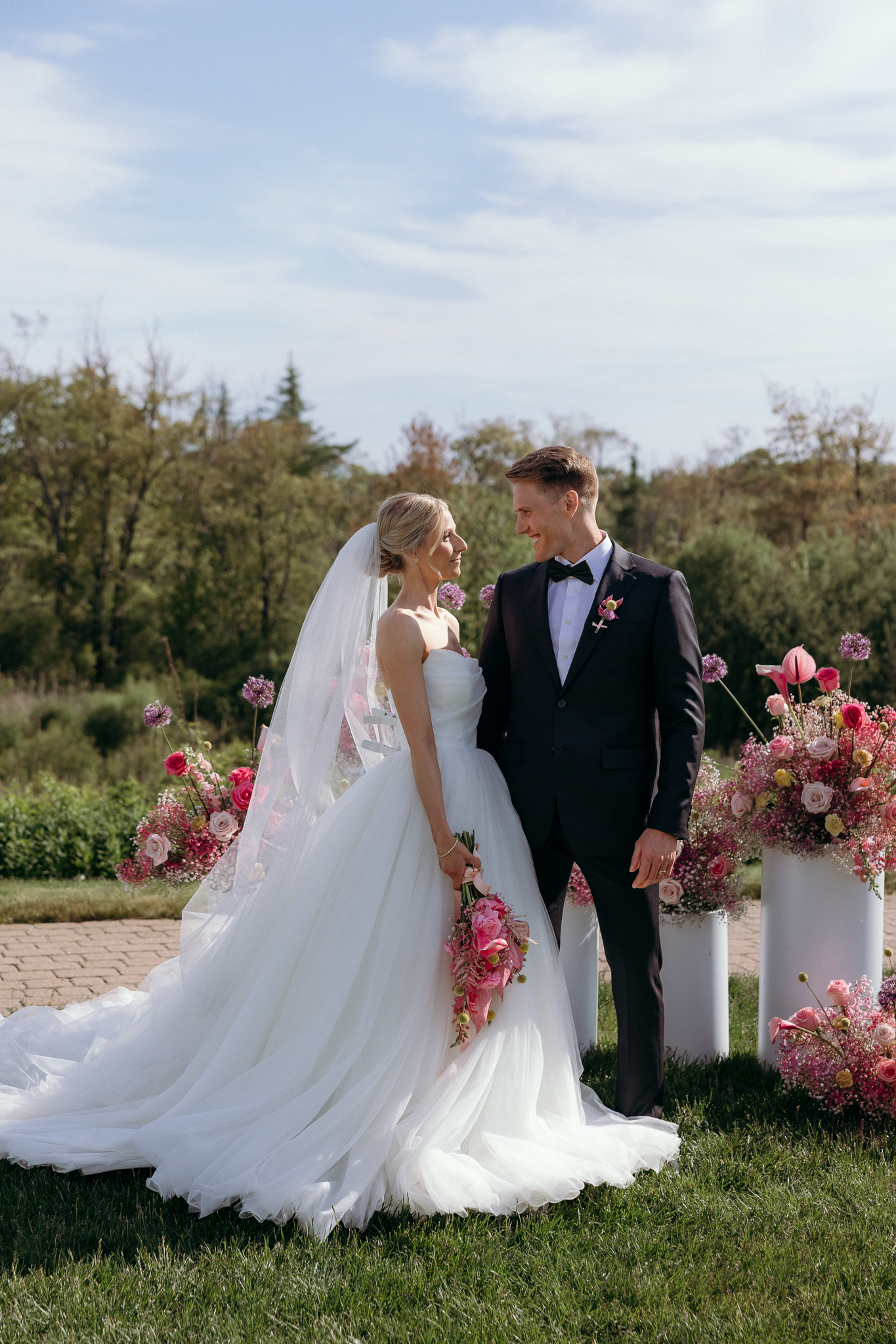 Inn by the Sea Maine | Sincerely Liz Photography Documentary and Editorial Wedding Photography | Bride in white gown and veil shares joyful moment with groom in black tuxedo. They stand outdoors with vibrant pink flowers, exuding happiness.