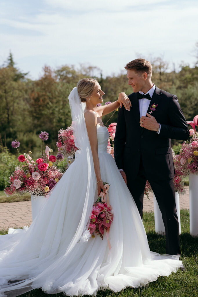 Inn by the Sea Maine | Sincerely Liz Photography Documentary and Editorial Wedding Photography | Bride in white gown and veil shares joyful moment with groom in black tuxedo. They stand outdoors with vibrant pink flowers, exuding happiness.