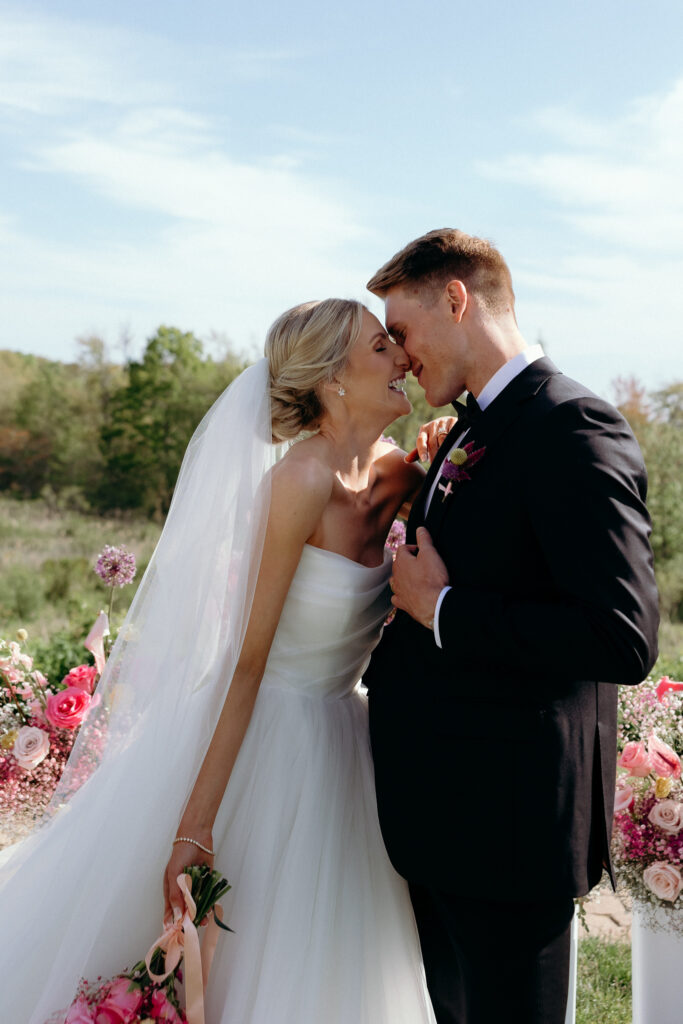 Inn by the Sea Maine | Sincerely Liz Photography Documentary and Editorial Wedding Photography | Bride and groom embracing outdoors, touching noses with eyes closed, smiling. Bride holds pink bouquet; groom wears black suit. Romantic setting.
