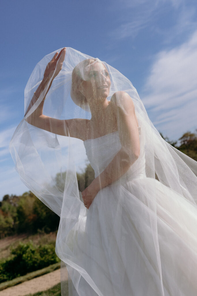 Inn by the Sea Maine | Sincerely Liz Photography Documentary and Editorial Wedding Photography | A bride in a strapless white gown joyfully lifts her veil against a clear blue sky. The scene is serene and filled with natural light.