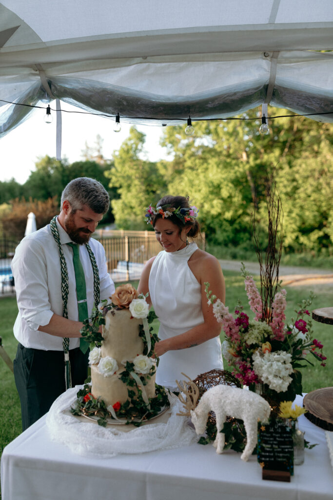 Vermont Wedding Photographer | Sincerely Liz Photography Documentary and Editorial Wedding Photography | Bride and groom cut a floral cake under a tent at an outdoor wedding. The bride wears a flower crown, and the groom dons a tie. Decor includes colorful blooms.