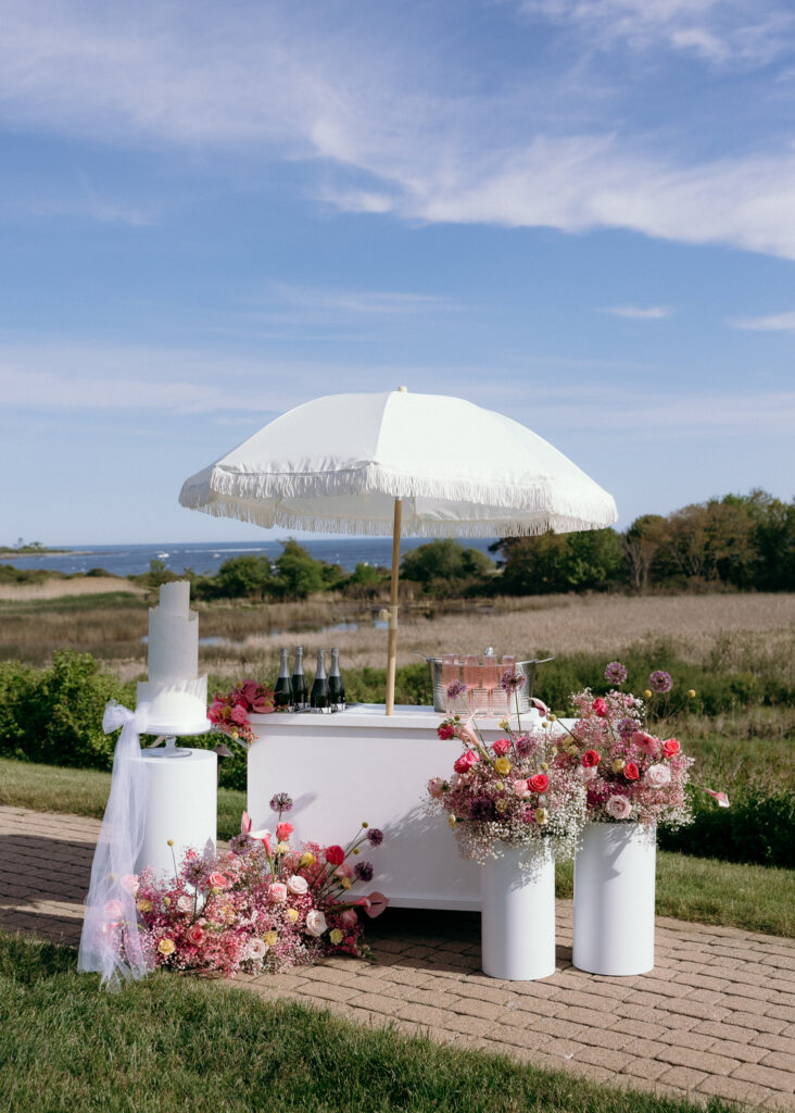Inn by the Sea Maine | Sincerely Liz Photography Documentary and Editorial Wedding Photography | Outdoor wedding setup with a white umbrella shading a bar. Vibrant floral arrangements and champagne bottles are displayed against a scenic field and sea backdrop.