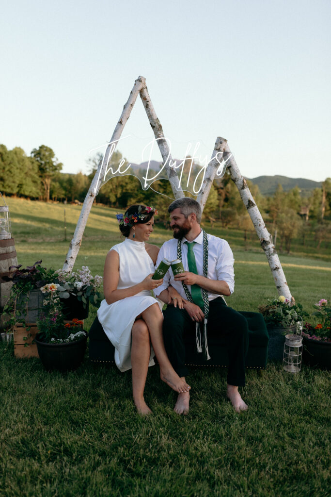Vermont Wedding Venues | Sincerely Liz Photography Documentary and Editorial Wedding Photography | A couple dressed in a white dress and green accents sits on a rustic bench in a scenic field. They are barefoot and smiling at each other, with "The Duffys" written above them. The backdrop features birch wood and flowers, conveying a joyful and serene wedding atmosphere.