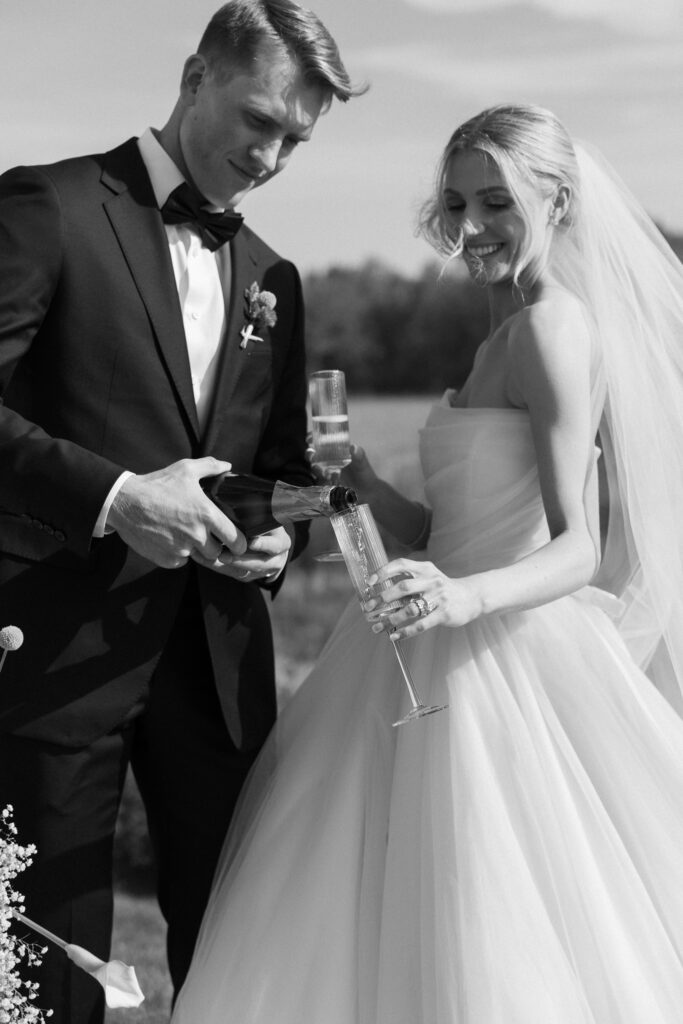 Vermont Wedding Venues | Sincerely Liz Photography Documentary and Editorial Wedding Photography | A bride in a flowing gown smiles as a groom in a tuxedo pours champagne into her glass. They stand outside on a sunny day, exuding joy and celebration.