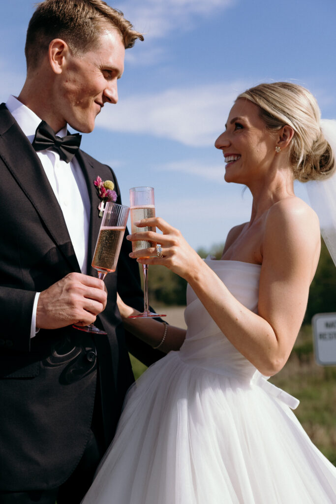 Vermont Wedding Photographer | Sincerely Liz Photography Documentary and Editorial Wedding Photography | A joyful couple in wedding attire toasts with champagne under a clear blue sky. The groom wears a tuxedo; the bride wears a strapless gown.