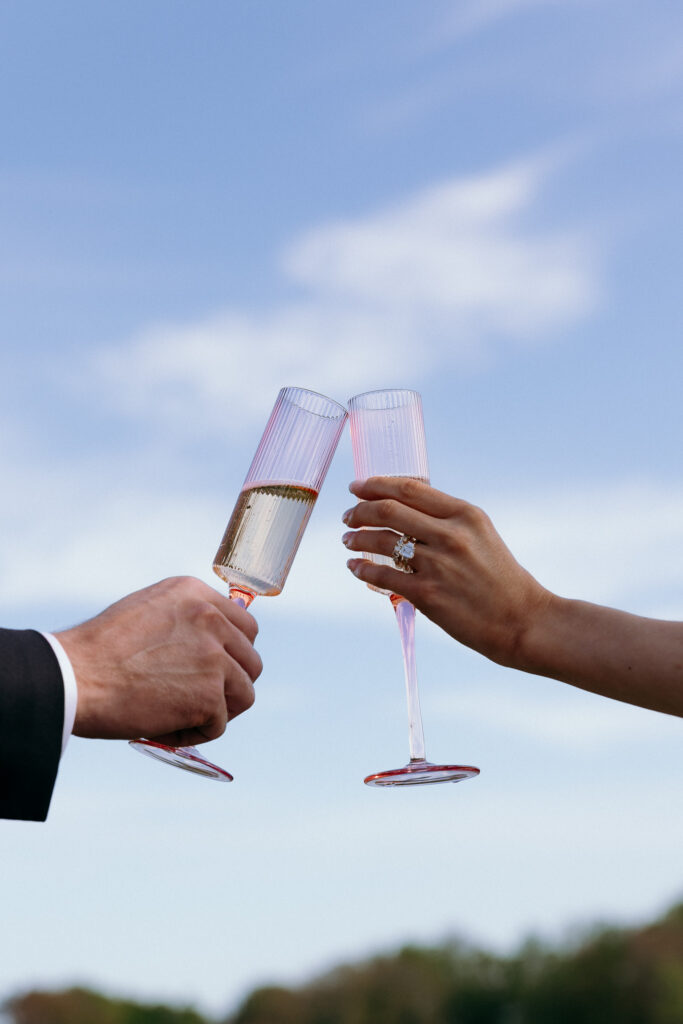Two hands raise pale pink champagne flutes against an open sky, a celebratory detail inspired by the atmosphere of VT wedding venues.