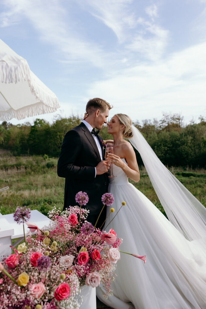 The couple stands outdoors holding champagne glasses beside a colorful floral arrangement, sharing a quiet celebratory moment at one of the top 10 wedding venues in Vermont.