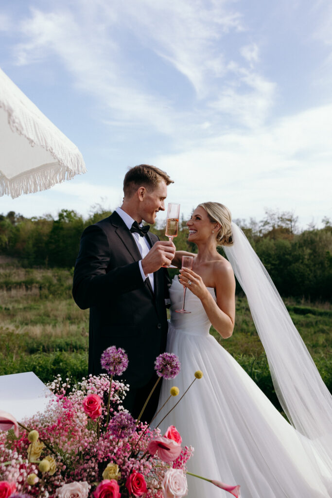 Bride and groom toast beneath open sky beside bright floral arrangements, an elegant outdoor portrait for small wedding venues in VT with scenic ceremony space.