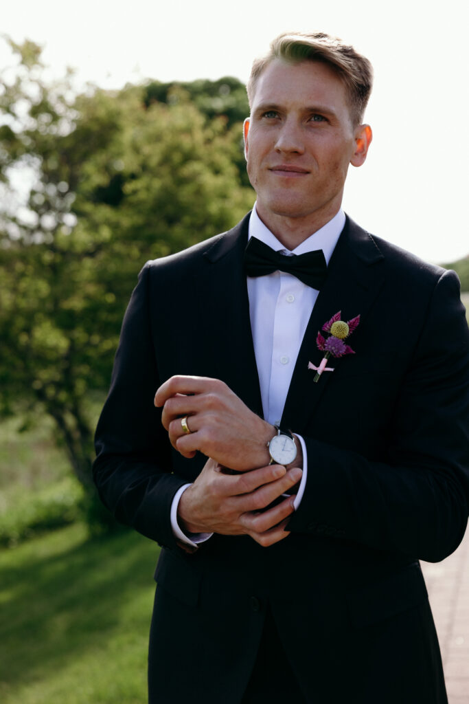 The groom stands in a black tuxedo outdoors, adjusting his cuff in afternoon light during a wedding in Vermont.