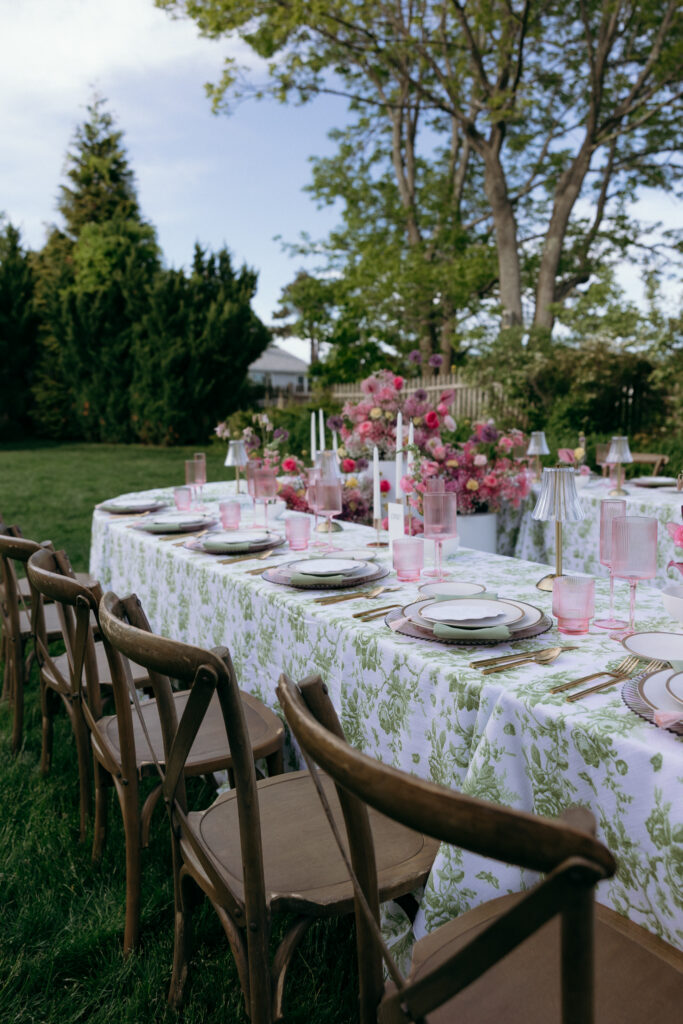 A long outdoor reception table is set with pink florals and patterned linens under the trees, a romantic look for small wedding venues in VT.